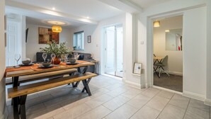 Ein Essbereich mit einem Holztisch, Bänken und einem Blumenstrauß. Im Hintergrund befindet sich eine Couch und ein Fenster.