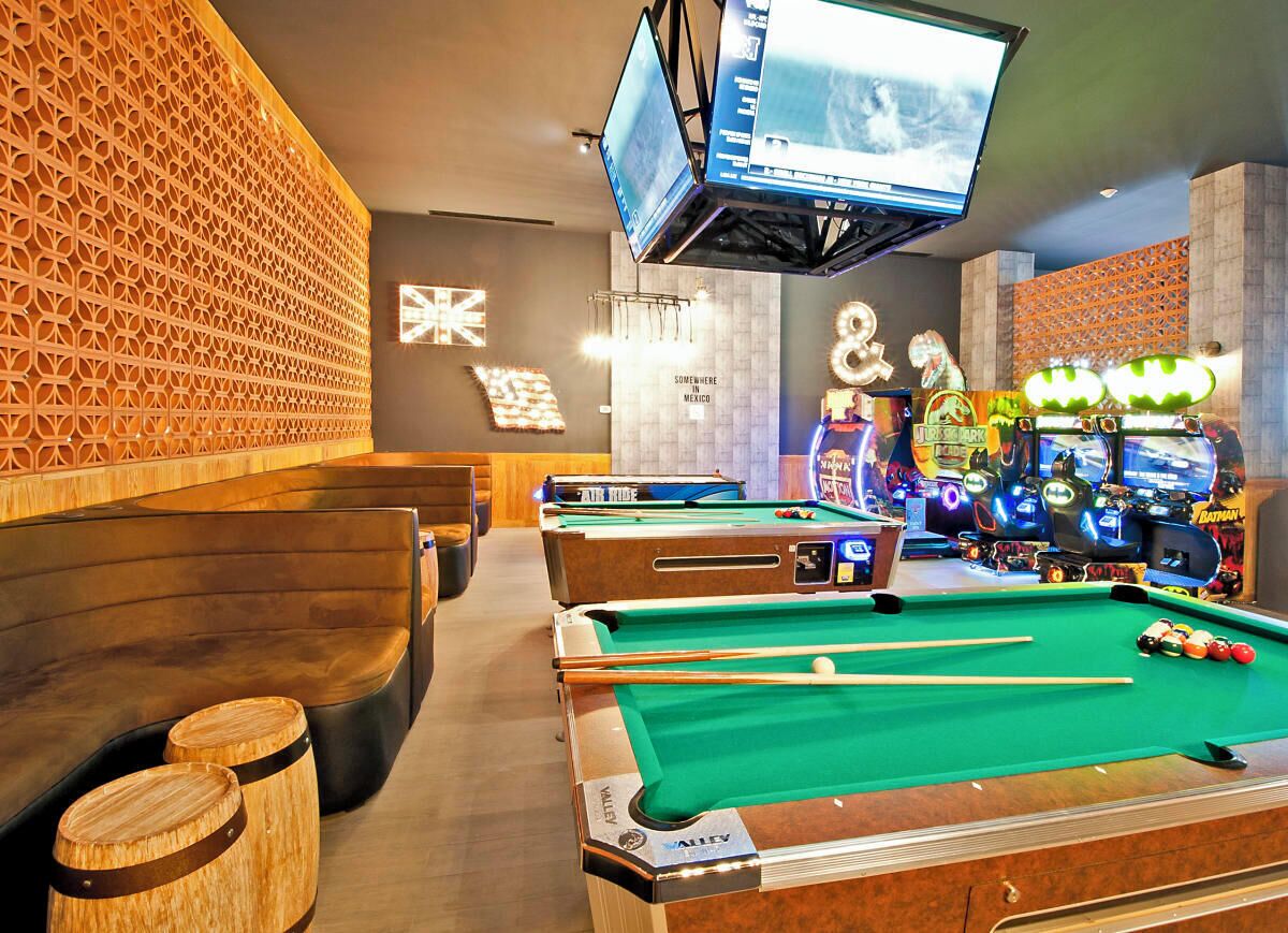 Un espace dédié aux tables de billard, avec plusieurs tables, un téléviseur fixé au plafond et, au fond, des jeux d’arcade.
