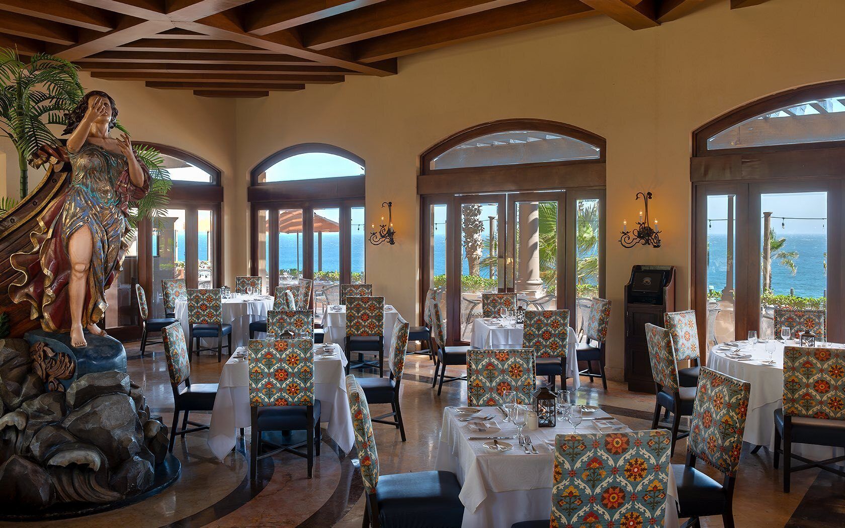 A restaurant with tables and chairs, a statue, and large windows with a view of the ocean.