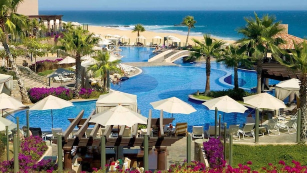 Zwembad van een resort met uitzicht op de oceaan, omringd door palmbomen en ligbedden.