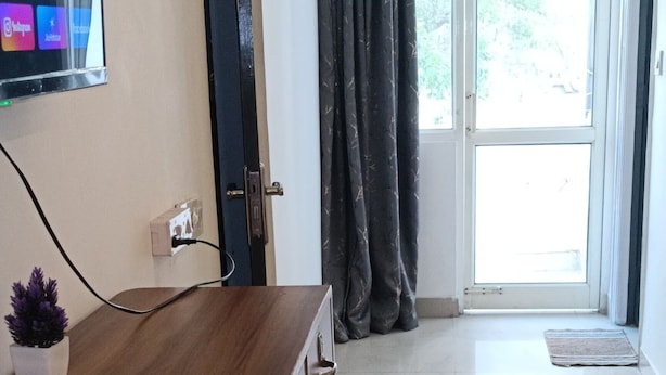 A room with a wooden desk, a television mounted on the wall, a door, and a large window with curtains.