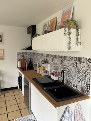 Une cuisine avec un évier noir, des armoires blanches et un dosseret à motifs. On y trouve des plantes suspendues et une étagère ornée d’objets décoratifs.