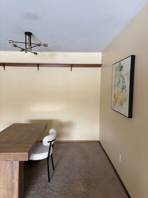 A room with a wooden table, a white chair, a ceiling light fixture, and a wall-mounted abstract painting.