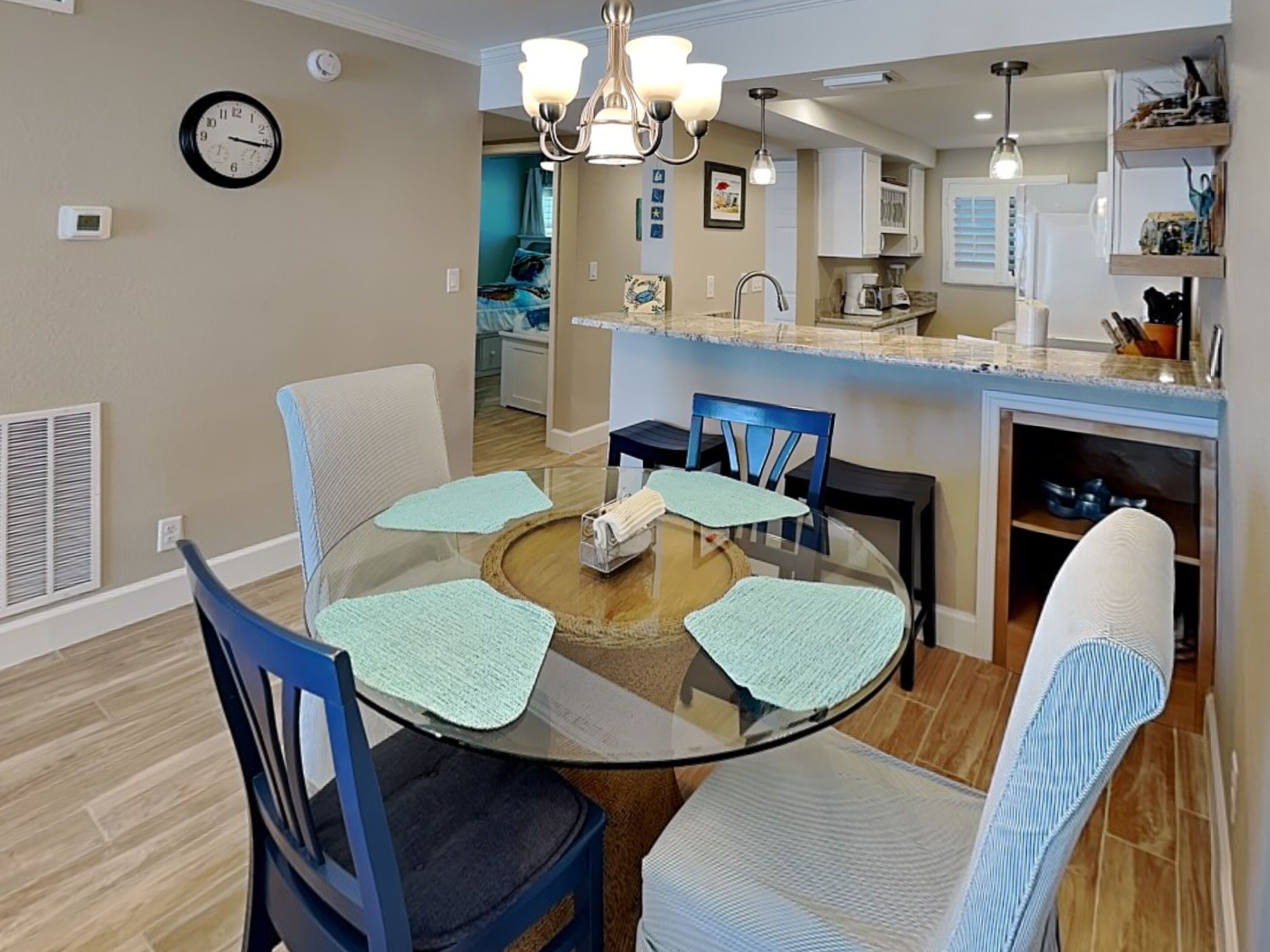 A dining area with a glass table surrounded by chairs, a kitchen in the background, and a clock on the wall.