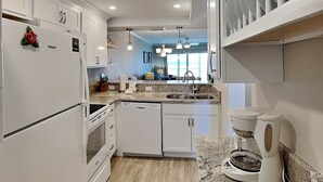 A modern kitchen with white cabinetry, a granite countertop, a built-in dishwasher, and a coffee maker. - Castle Beach 105 (Fort Myers Beach)