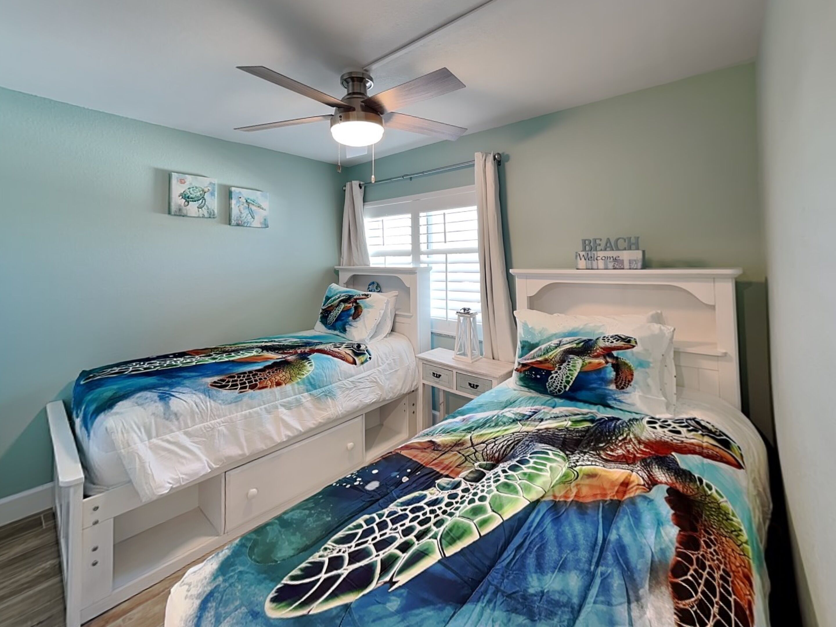 A bedroom with two beds, a ceiling fan, a window with curtains, and a 'BEACH Welcome' sign.