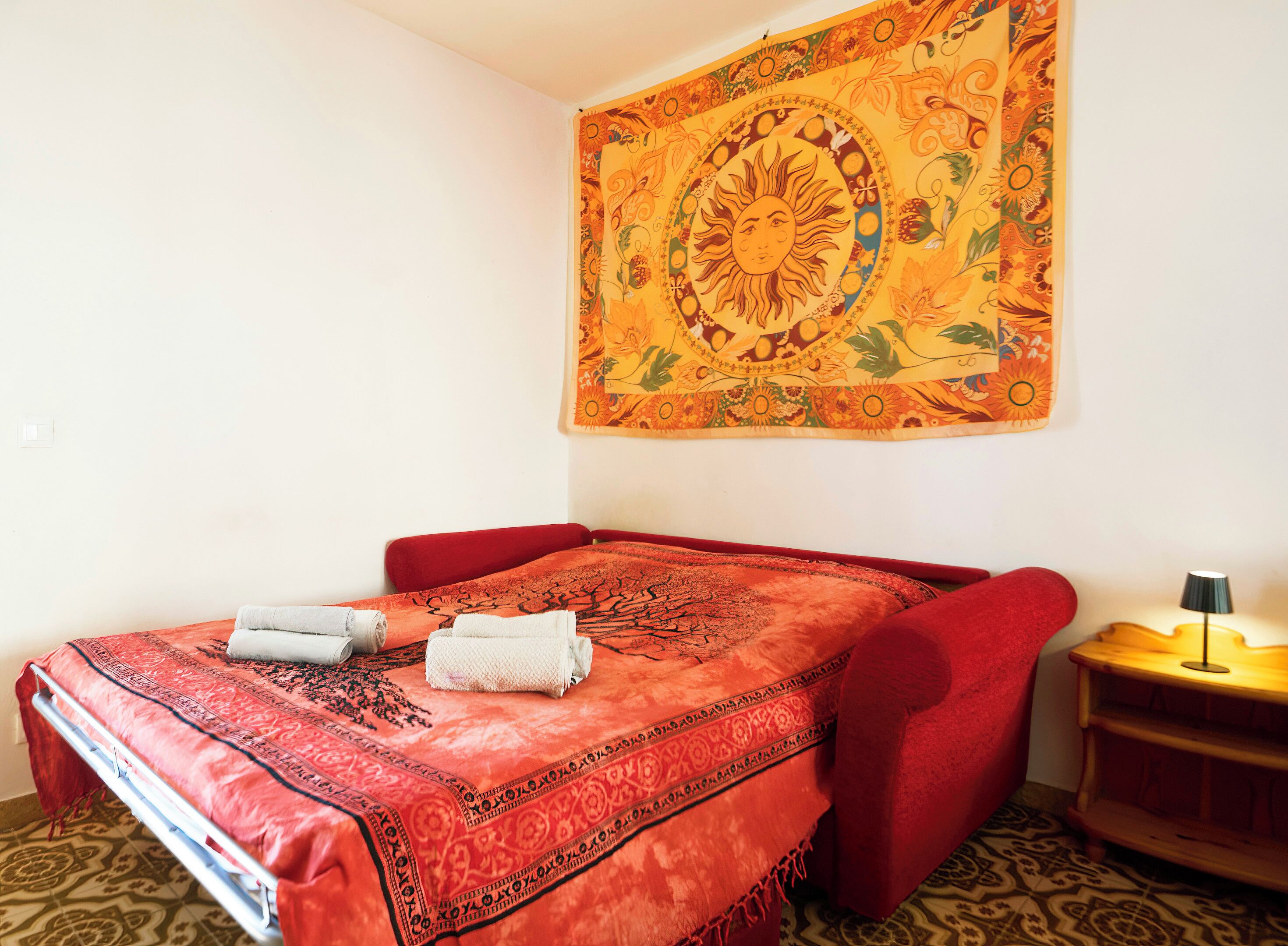 A room with a bed, a tapestry, a lamp, and a small wooden table.