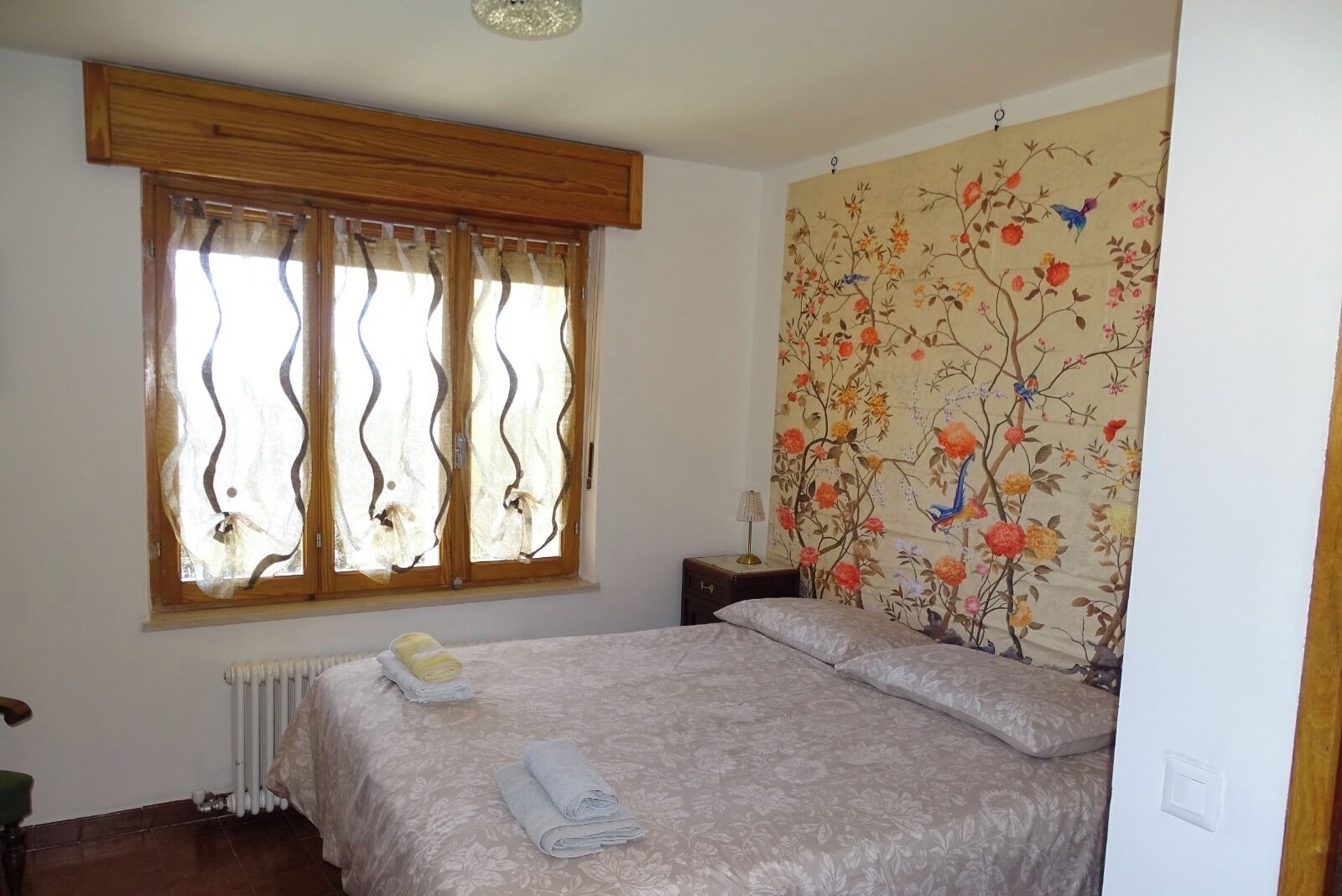 A bedroom with a bed, a radiator, a window with curtains, and a floral wallpaper.