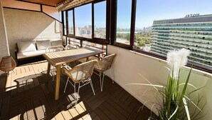 Un balcón con mesa y sillas de madera, un sofá y un ventanal amplio con vista a la ciudad.