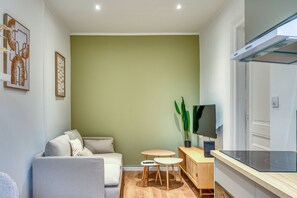 A modern living room with a sofa, a small table, a TV, and a kitchen area with a stove. - Le Zola – Oullins – 10 min à pied métro B – Wi-Fi inclus (Oullins-Pierre-Bénite)
