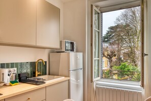 A kitchen with a white refrigerator, a microwave, a sink with a gold faucet, and a window with a view of trees and buildings. - Le Zola – Oullins – 10 min à pied métro B – Wi-Fi inclus (Oullins-Pierre-Bénite)