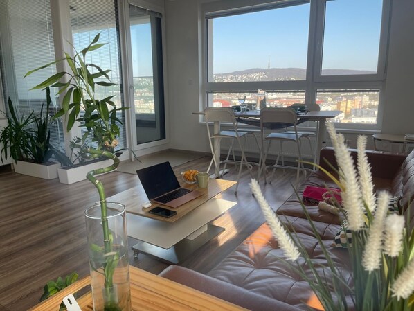 A modern living room with a laptop on a table, a sofa, and large windows offering a city view.
