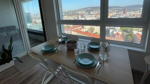 Un coin repas avec une table dressée pour quatre personnes, de grandes fenêtres offrant une vue sur la ville et une plante en pot placée sur la gauche.