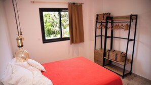 A bedroom with a red bedspread, a hanging lamp, a window with a view of greenery, and a wooden shelf with baskets and hangers. - VILLA NEPITA Modern 1-bedroom with private pool (Porto-Vecchio)