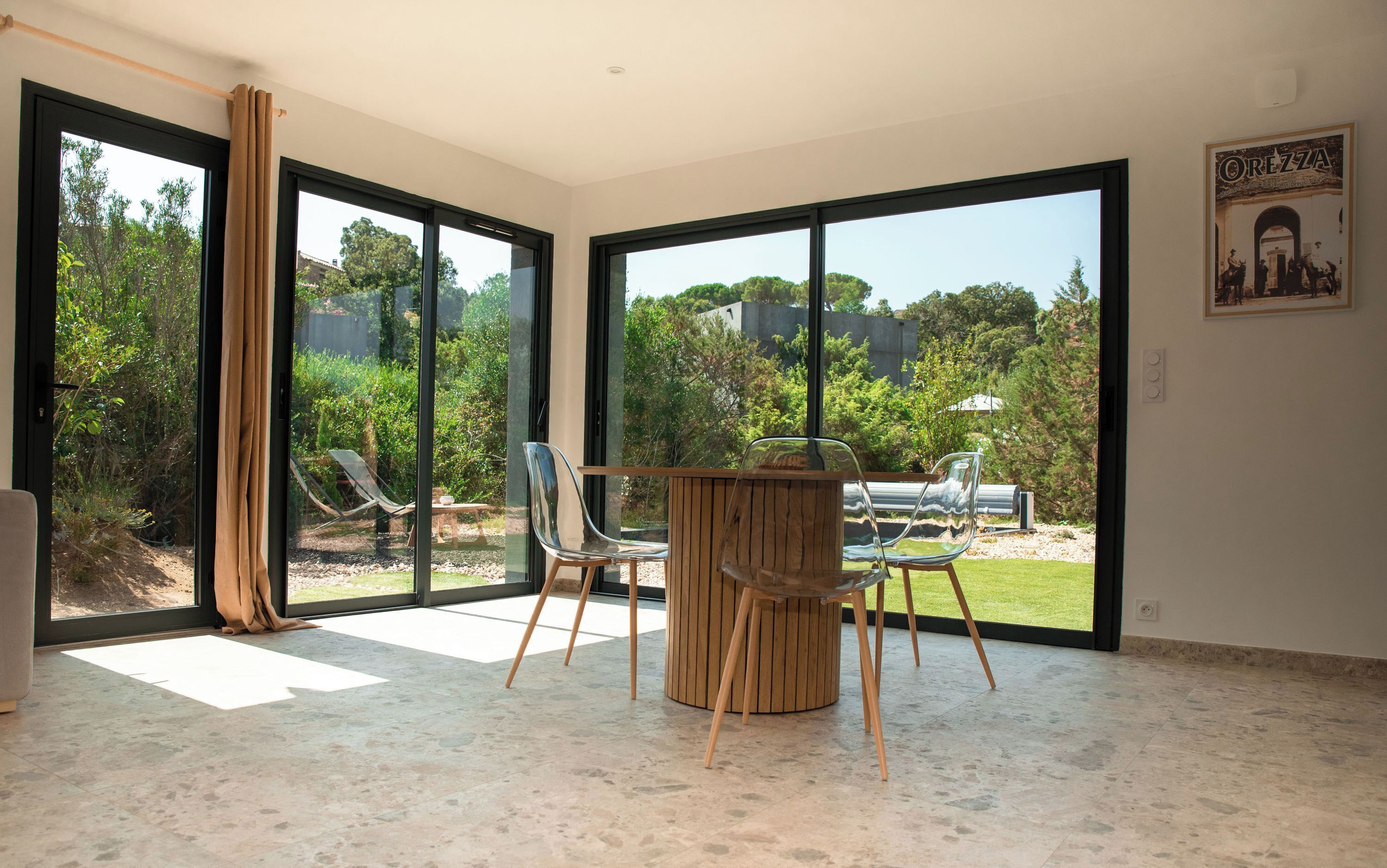 A spacious room with large sliding glass doors leading to a garden, a wooden table with chairs, and a poster on the wall.