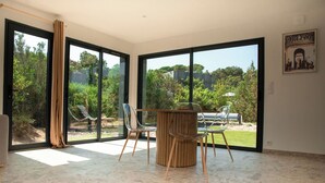 A spacious room with large sliding glass doors leading to a garden, a wooden table with chairs, and a poster on the wall. - VILLA NEPITA Modern 1-bedroom with private pool (Porto-Vecchio)