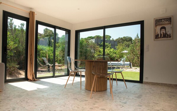 A spacious room with large sliding glass doors leading to a garden, a wooden table with chairs, and a poster on the wall. - VILLA NEPITA Modern 1-bedroom with private pool (Porto-Vecchio)