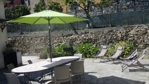 Eine Terrasse im Freien mit Tisch und Stühlen, einem grünen Sonnenschirm und einer Steinsäule im Hintergrund.