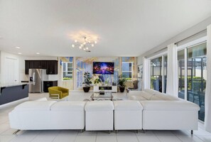 A modern living room with a white sectional sofa, a yellow chair, a flat-screen TV, and a dining area with a kitchen in the background. - 9BR The Sunset Paradise Resort w/ Pool Near Disney (Kissimmee)