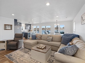 A modern living room with a sectional sofa, a coffee table, and a gray armchair. There is a kitchen area in the background with a view of the outdoors. - Hot tub, Waterfront, Sunset Views, Dock, Lilypad, The Blue Blessing (Goreville)