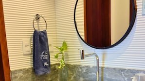 Un baño con un espejo redondo, un lavabo blanco, un toallero con una toalla azul, una planta pequeña y un estante de madera con artículos de aseo.