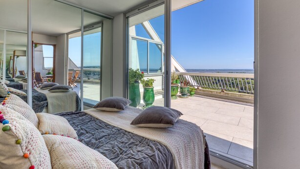 Ein modernes Schlafzimmer mit einem großen Bett, einem Balkon mit Blumentöpfen und Blick auf das Meer.