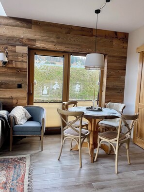 A cozy room with a wooden wall, a round wooden table with chairs, a blue sofa, and a large window with a view of greenery. - Izera Polana Also bookable as 2-3 houses (Świeradów-Zdrój)