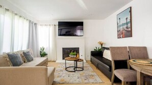 Una sala de estar moderna con un sofá, una mesa de centro y una chimenea. Hay un cuadro en la pared y una planta en el suelo.