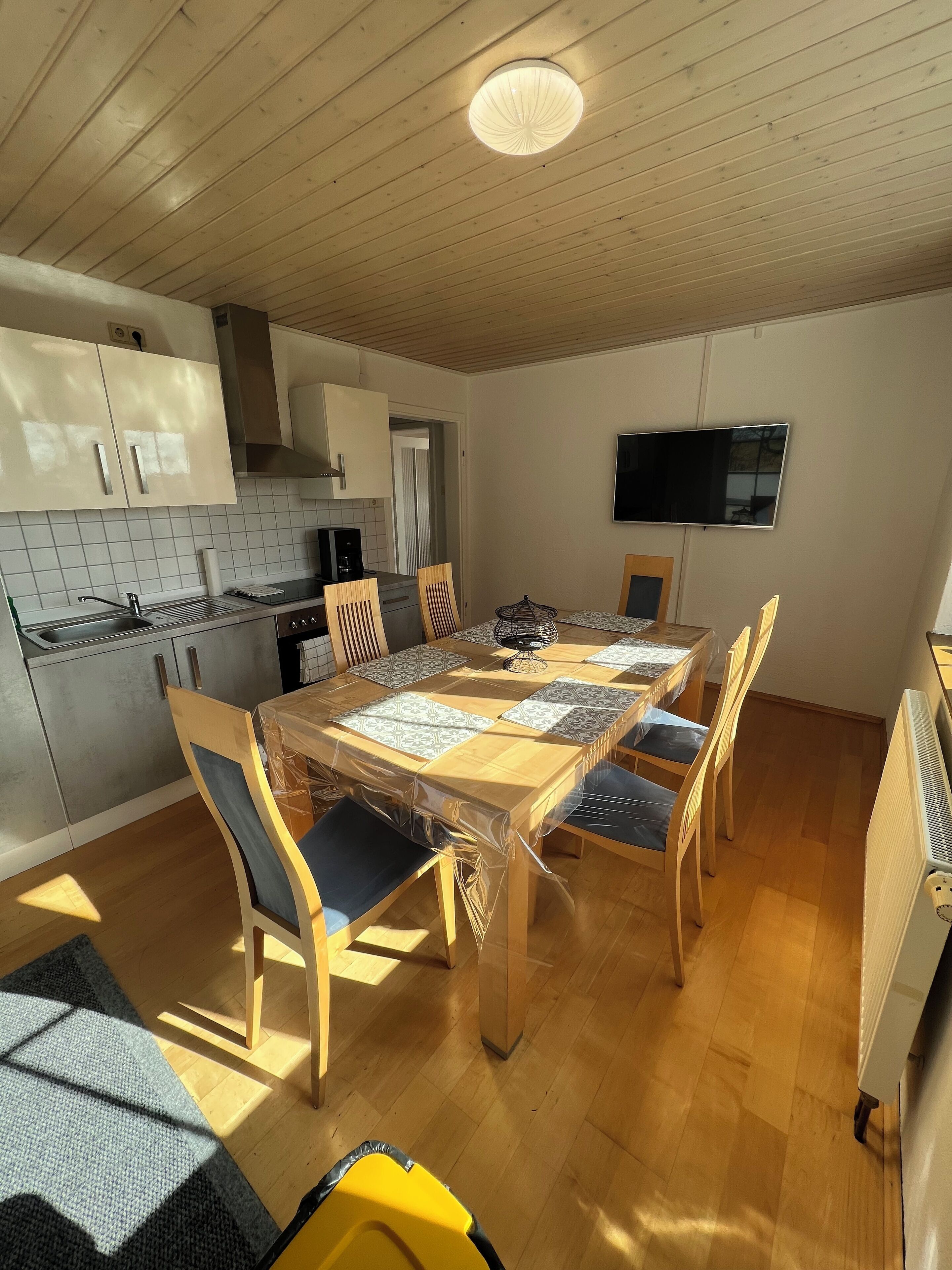 Un comedor con mesa y sillas de madera, una cocina con armarios blancos y un salpicadero de azulejos, y un televisor de pantalla plana montado en la pared.