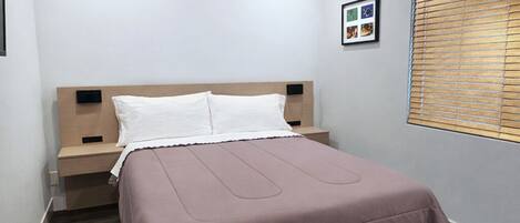 Ein Schlafzimmer mit einem Bett, einem Nachttisch, einem Wandbild, einem Fenster mit Jalousien und einem Deckenventil.