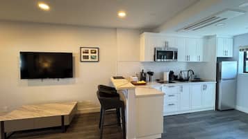 Een moderne keuken met witte kasten, een magnetron, een koelkast, een eethoek met een bank en een flatscreen-televisie aan de muur.