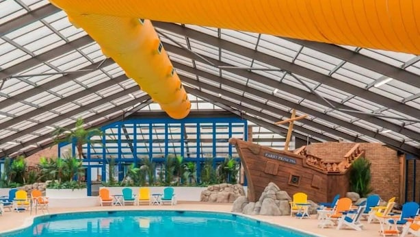 An indoor swimming pool with a clear water surface, surrounded by lounge chairs and a wooden structure resembling a shipwreck. - North Wales Coastal Stay (Rhyl)
