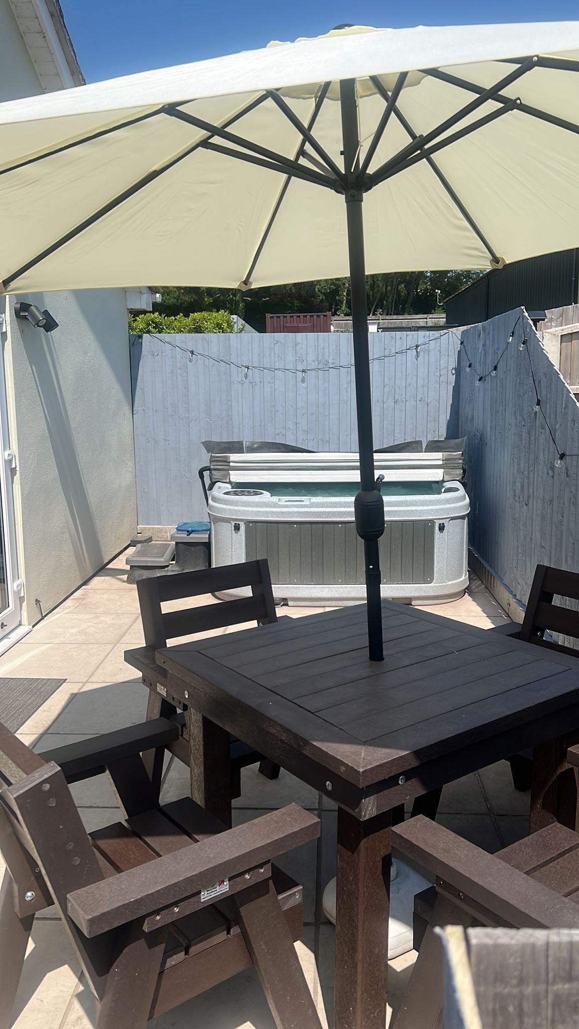 Un espace extérieur aménagé en terrasse, avec une table et des chaises en bois, un grand parasol et un jacuzzi en arrière-plan.