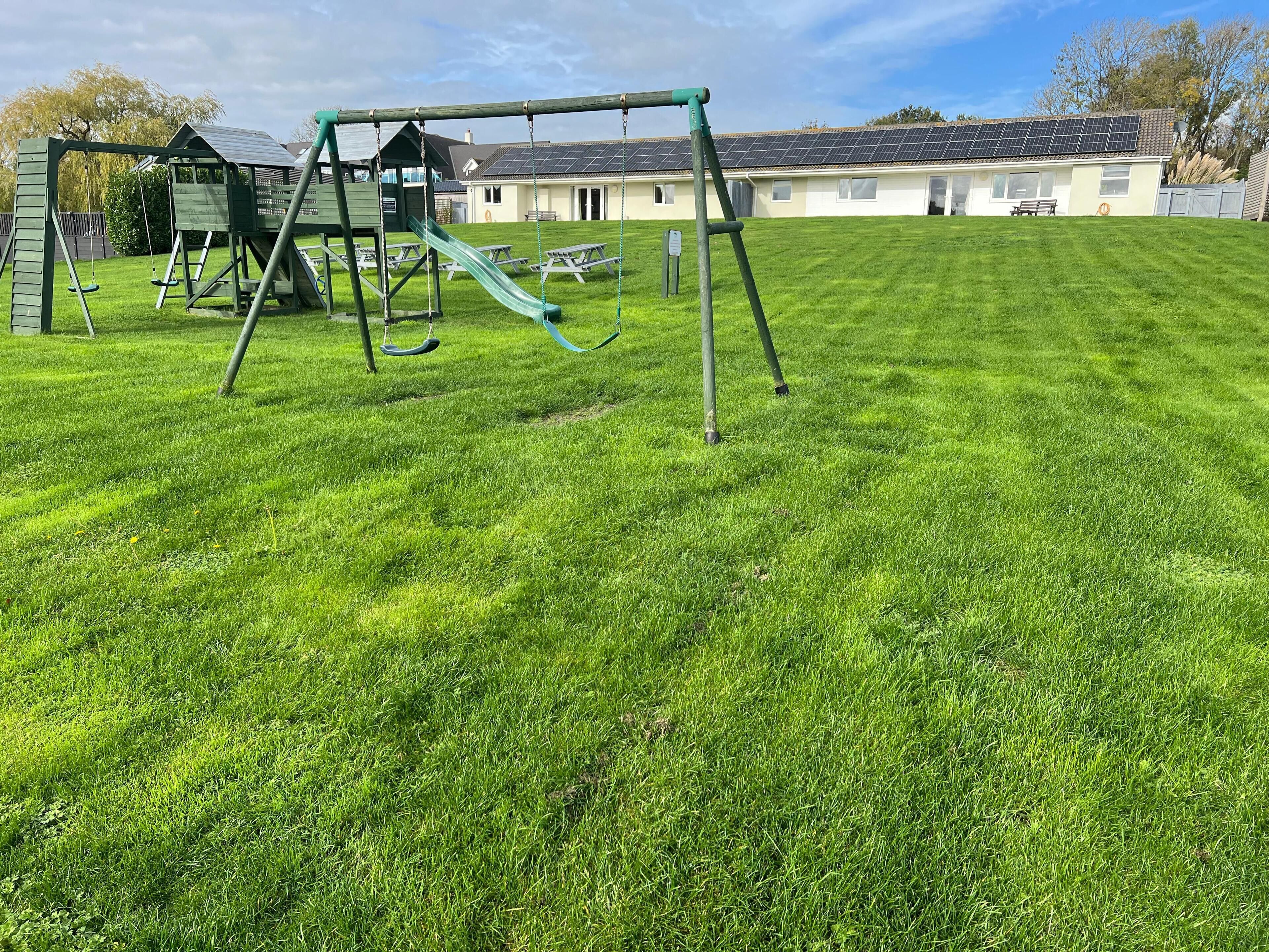 Une aire de jeux avec de l’herbe, des balançoires et un toboggan, une table de pique-nique, et un bâtiment doté de panneaux solaires en arrière-plan.