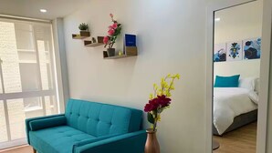 A modern room with a teal sofa, a small round table with a vase of flowers, a wall-mounted shelf with decorative items, and a large mirror reflecting a bed and a wall art piece. - Lovely loft downtown - La Candelaria (Bogotá)