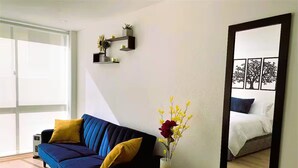 Un salon moderne avec un canapé bleu, des coussins jaunes, une petite table ronde ornée d’un vase de fleurs et un grand miroir accroché au mur.