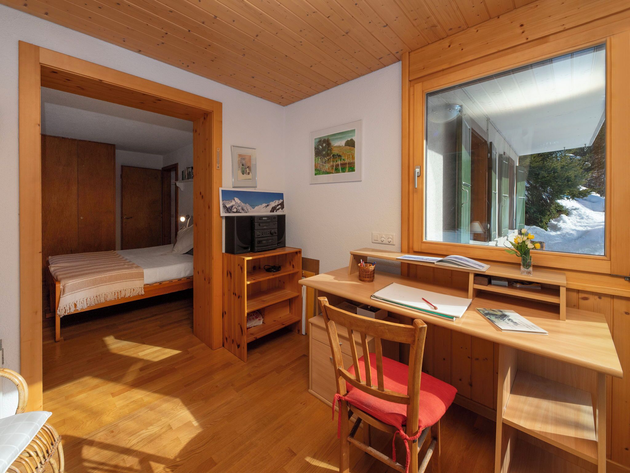 A cozy room with a wooden ceiling, a bed, a desk with a chair, a TV, and a window with a snowy view.
