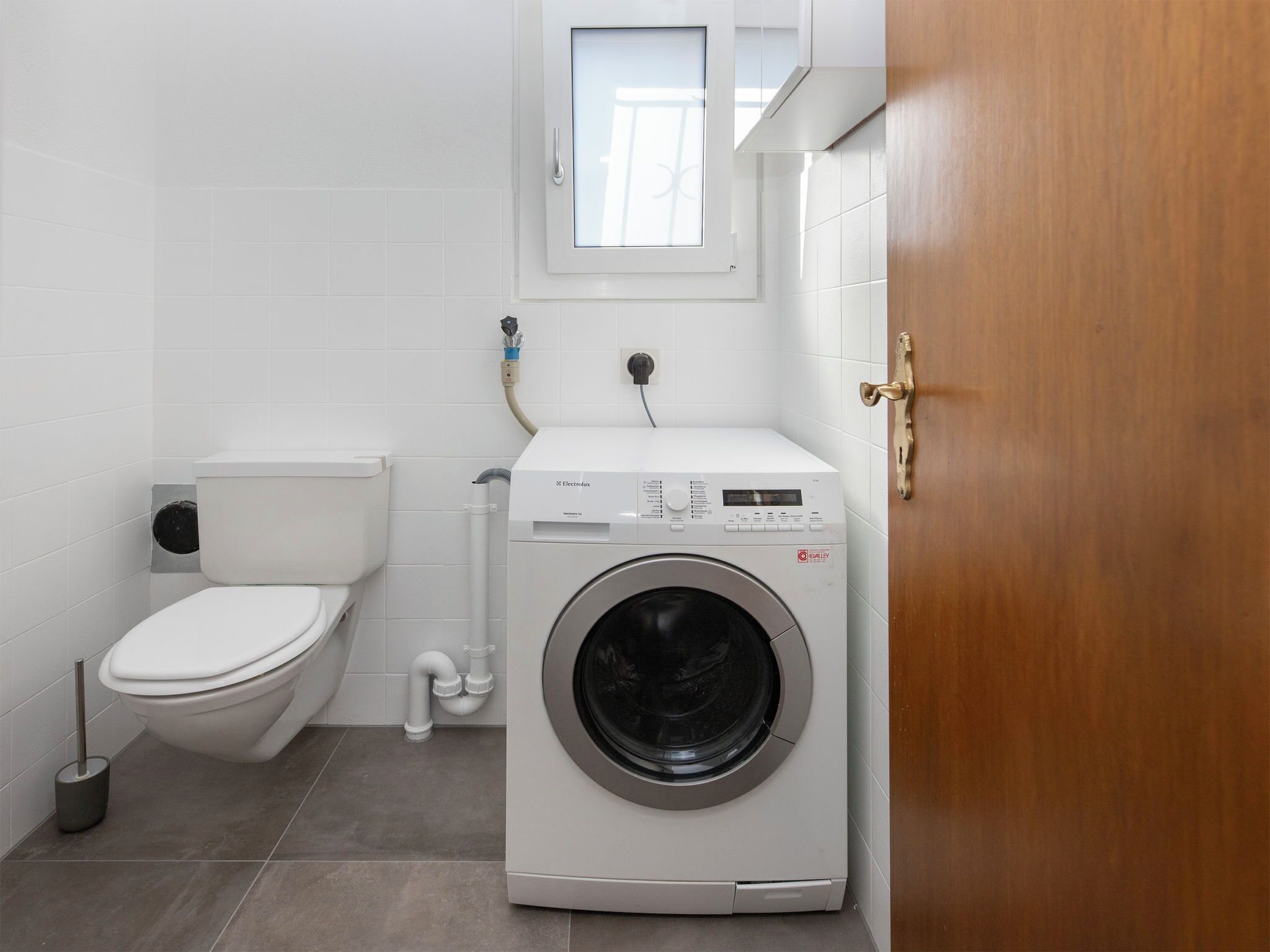 A compact bathroom with a toilet, washing machine, and a window above the sink.
