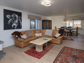 Ein Wohnzimmer mit braunem Ledersofa, einem Couchtisch und einem gemusterten Teppich. An der Wand hängt ein Gemälde und es gibt eine Essecke mit Tisch und Stühlen.