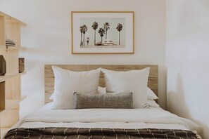 A neatly made bed with white linens and a brown striped blanket, a wooden headboard, and a framed picture of palm trees and a car on the wall.