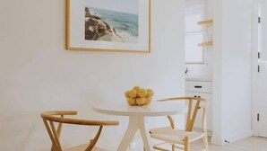Ett minimalistiskt rum med ett vitt bord och två trästolar, en inramad tavla på väggen och en vit dörr i bakgrunden.