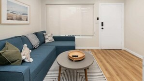 Una sala de estar con un sofá azul, una mesa redonda para café, un cuadro en la pared, una ventana con persianas y una puerta blanca.