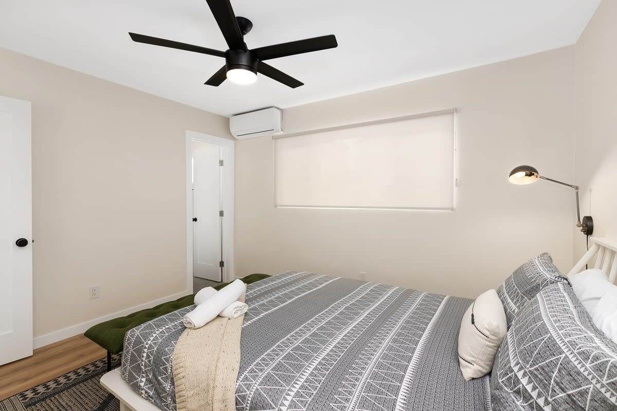 Ein Schlafzimmer mit Bett, Deckenventilator, Fenster mit Jalousien, Nachttischlampe und einer Tür.