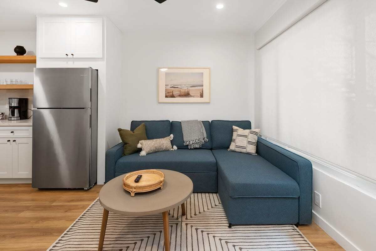A modern living room with a blue sectional sofa, a small round table, and a kitchen area with a stainless steel refrigerator.