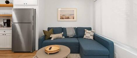 A modern living room with a blue sectional sofa, a small round table, and a kitchen area with a stainless steel refrigerator.