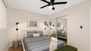 Un dormitorio con cama, un banco verde, ventilador de techo, espejo y mesita de noche.