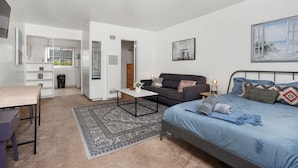 A modern, well-lit room with a bed, a sofa, a coffee table, and a desk. There is a kitchen area in the background. - Steps Away from PB Beaches + Hot Tub (San Diego)