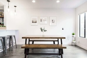 A dining area with a wooden table and bench, a white wall with framed pictures, and a window on the right. - Ocean Beach Retreat | 3BR Spanish‑Style Home - Steps to Santa Cruz Cove (San Diego)