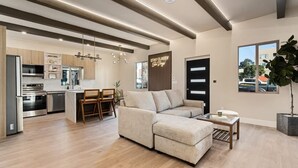 Une cuisine moderne dotée d’appareils électroménagers intégrés, un coin repas avec une table et des chaises en bois, un salon avec un canapé et une table basse, et une grande fenêtre offrant une vue sur l’extérieur.