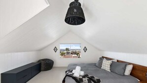 Quarto com teto inclinado, cama com roupa de cama cinza, luminária pendente preta, janela com vista para edifícios e um banco preto.